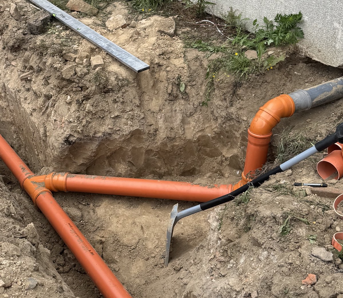 Wykop przy ścianie budynku z ułożoną pomarańczową rurą kanalizacyjną, trójnikiem i kolanem wchodzącym do budynku