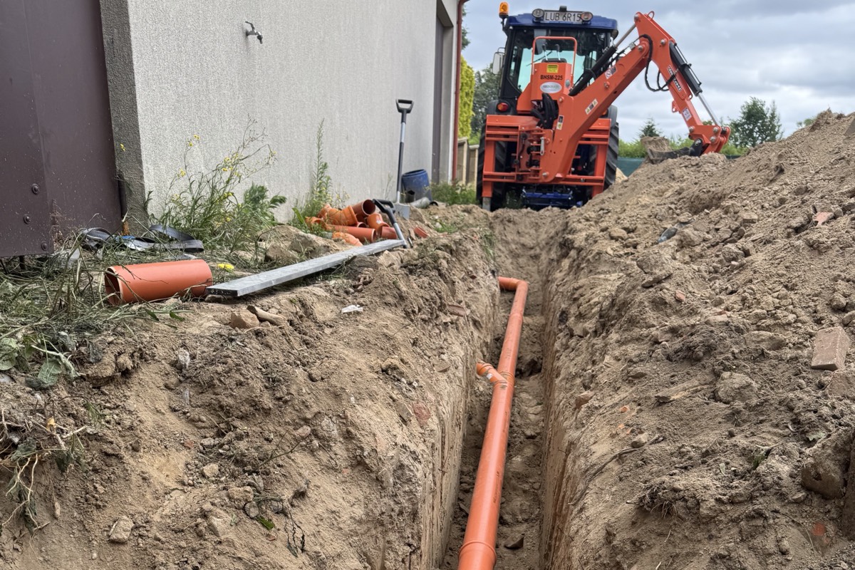 Wąski wykop pod kanalizację wzdłuż ściany budynku, na dnie ułożona pomarańczowa rura, w głębi koparka BHMS 225 na ciągniku Farmtrac