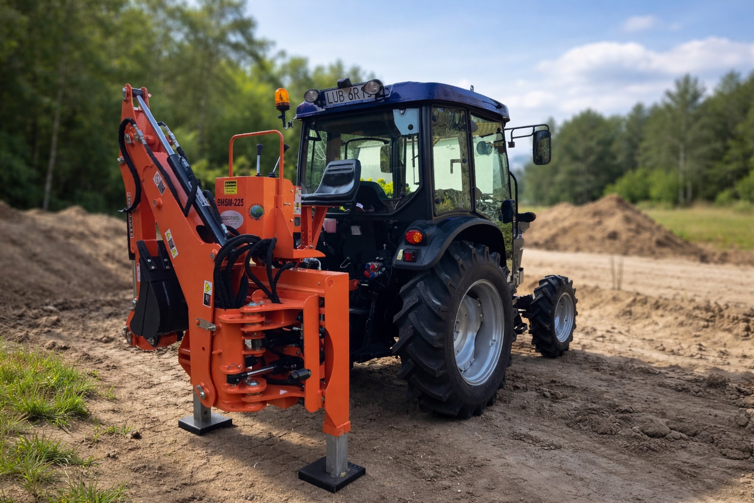 Ciągnik Farmtrac z zamontowaną z tyłu pomarańczową koparką BHMS 225, na piaszczystym placu przy pryzmach ziemi